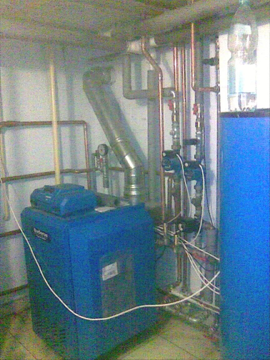 Niebieski piec CO marki Buderus w kotłowni z widocznymi rurami miedzianymi i białymi kablami.